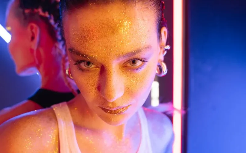 Close-up portrait of a woman with glitter makeup, standing under vibrant neon lights with earrings visible.