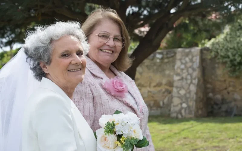 Two senior women in wedding attire smiling outdoors, holding a bouquet in a garden setting.