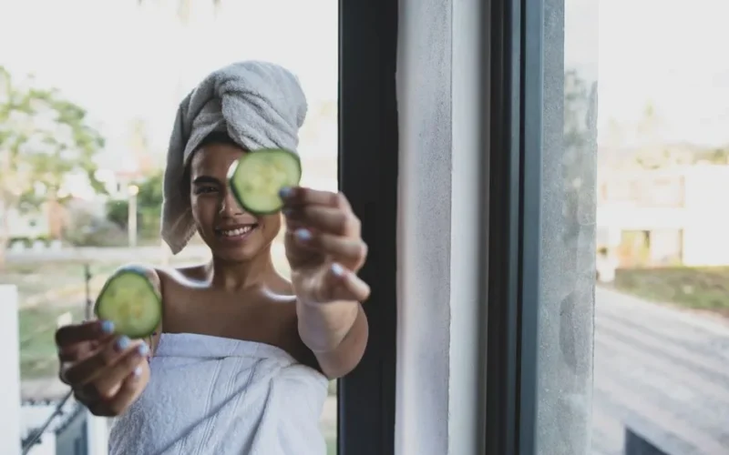 Smiling woman with towel turban holding cucumber slices, enjoying a skincare routine by the window.