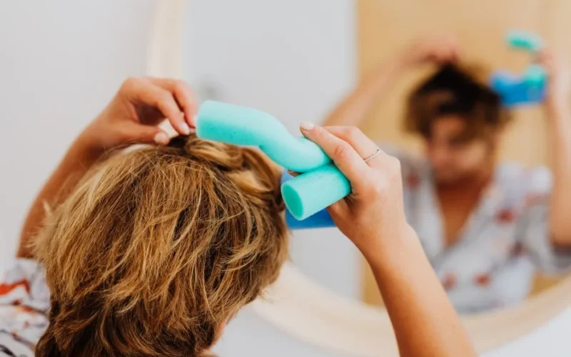 A woman using a curling rod to style her short hair while looking in a mirror.