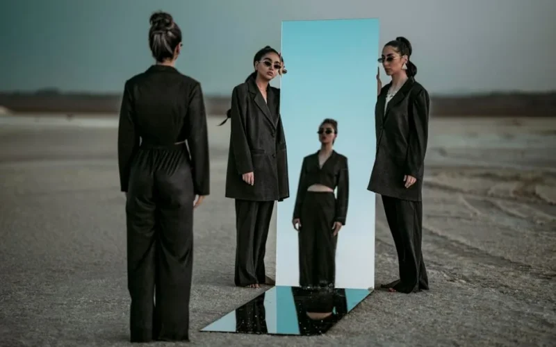 Elegant woman in black clothing reflects in a mirror placed outdoors, creating a surreal fashion scene.