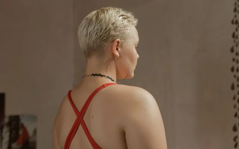 Stylish portrait showing a woman with short blond hair, large earrings, and a red top viewed from behind.