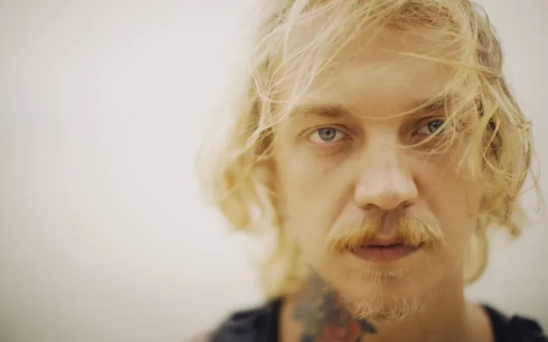 An artistic close-up portrait of a blonde man with facial hair and tattoos, conveying a modern, edgy style.