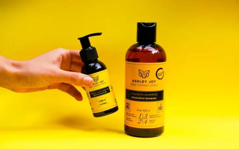 Hand holding a bottle of herbal hair care oil next to shampoo on yellow backdrop.