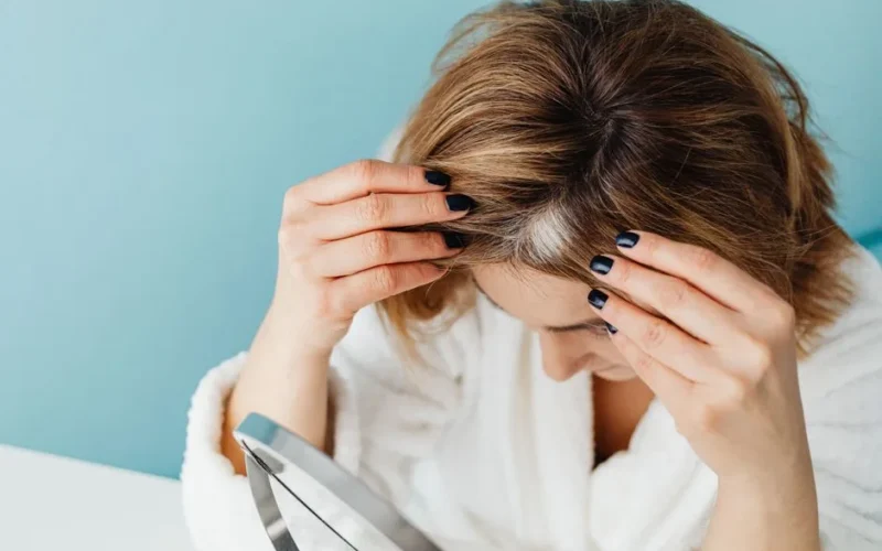 Woman in white robe checking her hair for gray strands using a mirror on blue background.