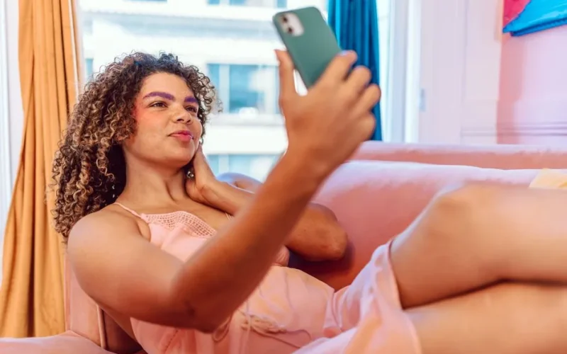 Woman with curly hair taking a selfie with smartphone while lounging on a comfortable indoor couch.
