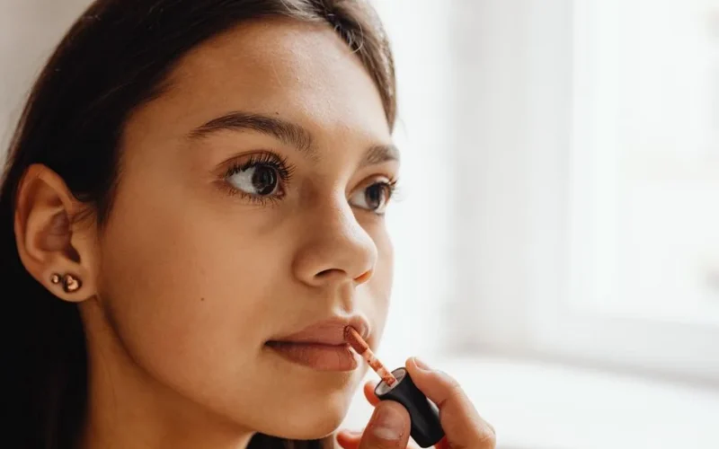 Close-up of a young woman applying lipstick indoors with natural daylight.