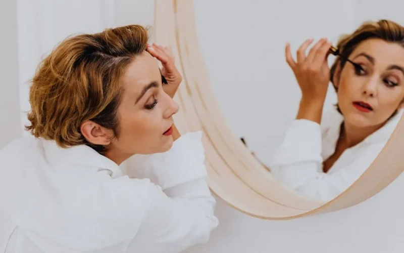 Woman applying mascara in a mirror, showcasing elegance and beauty in a candid indoor setting.