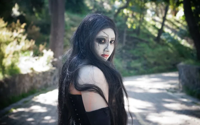 A woman with gothic makeup stands in a sunlit park, casting a mysterious look.