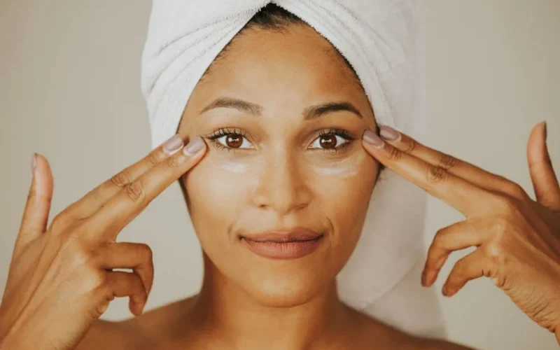 Woman applying facial cream with a towel wrapped on her head, highlighting skincare routine.