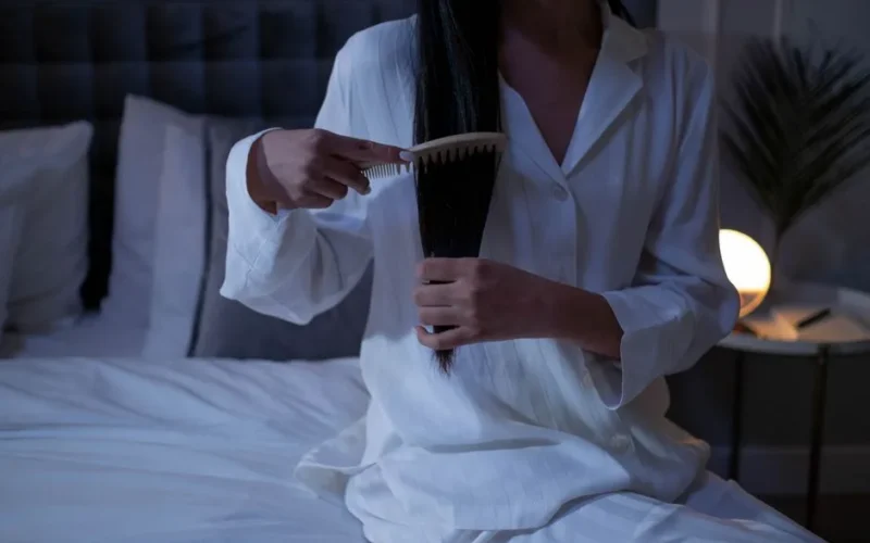 Woman combing long hair in a tranquil bedroom, dressed in pajamas.