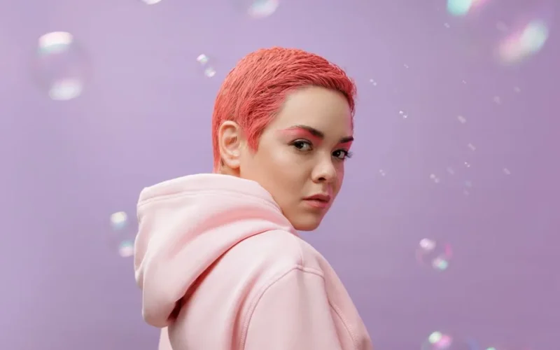 Vibrant studio portrait of a woman with pink hair wearing a hoodie against a lilac background.