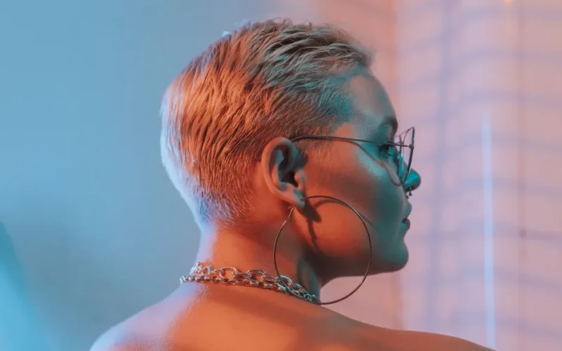 Close-up profile of a stylish woman with blonde short hair, hoop earrings, and chain necklace in vibrant lighting.