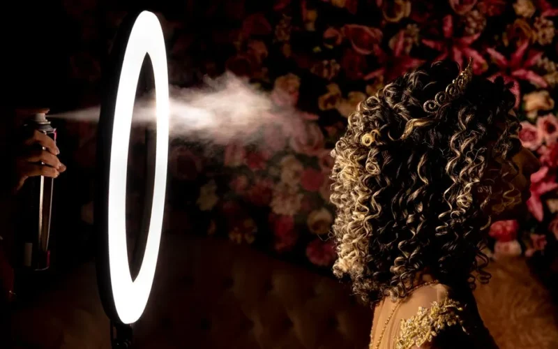 Side profile of a woman with curly hair being styled with hairspray under a ring light, perfect for beauty themes.