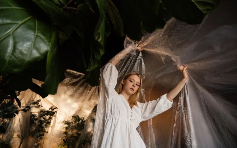 Artistic composition of a woman in a white dress surrounded by plastic and foliage.
