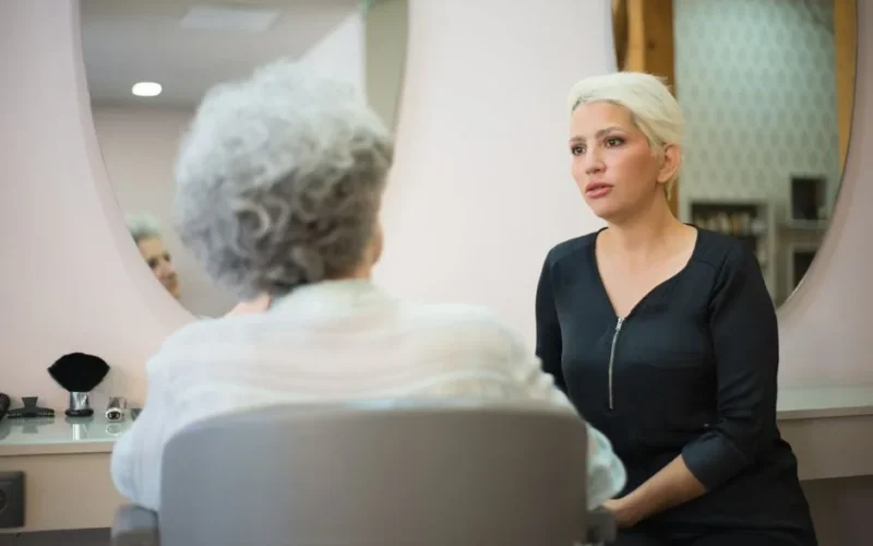 Two women having a conversation in a salon setting with a mirror reflecting their interaction.