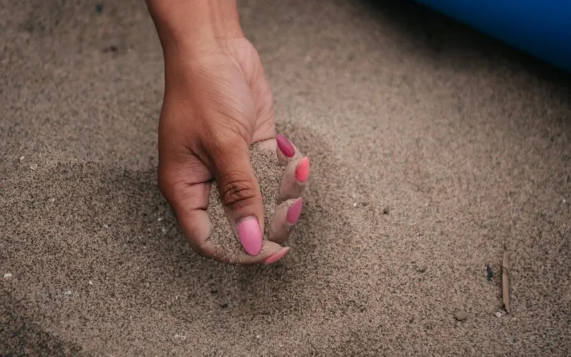 A close-up photo of a hand with pink manicured nails holding sand on a beach.