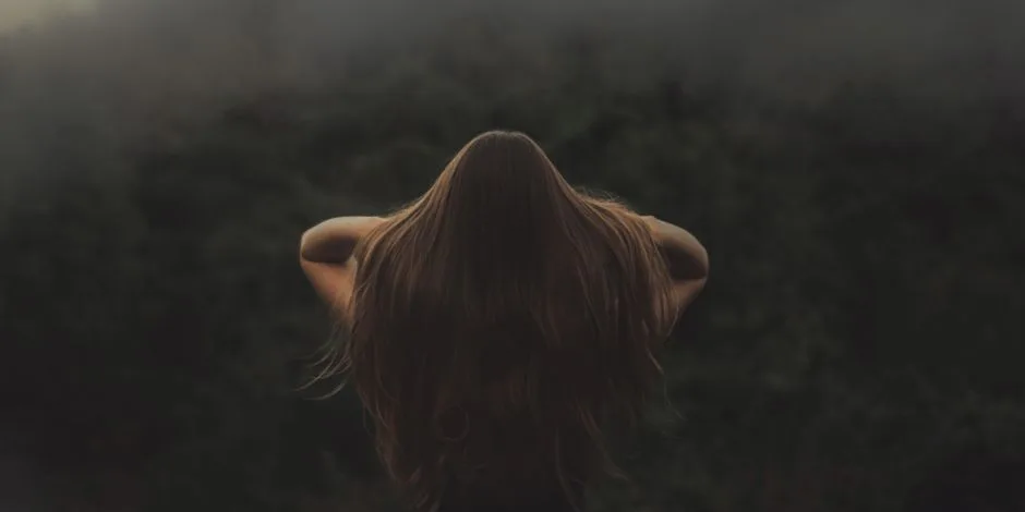 Back view of a woman with long hair, standing in a misty forest.