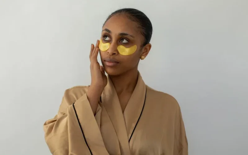 Woman wearing gold under-eye patches in a robe, promoting skincare and relaxation.