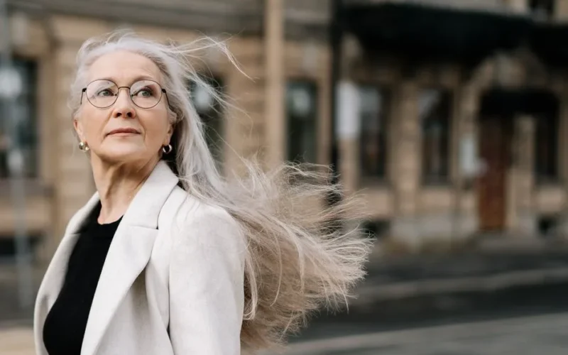 Stylish senior woman with flowing hair and eyeglasses walking in a breezy urban environment.