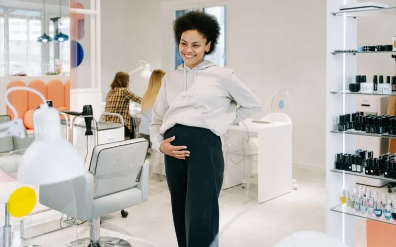A smiling pregnant woman stands in a stylish nail salon, showcasing joy and relaxation.