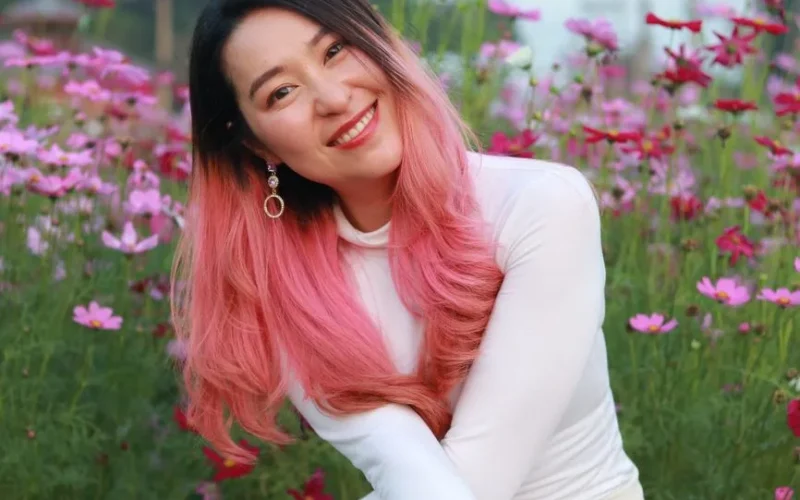 A cheerful woman with pink ombre hair smiles amidst vibrant flowers.
