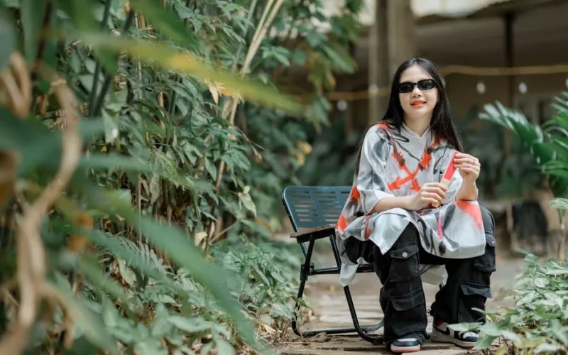Stylish woman wearing sunglasses and oversized hoodie sitting in a garden surrounded by plants.