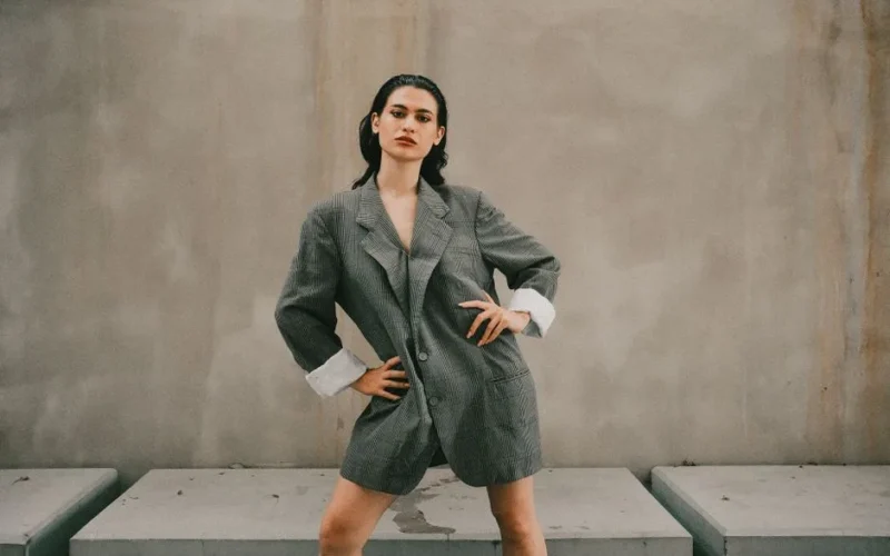 Stylish woman poses confidently in an oversized grey suit against a minimalist outdoor backdrop.