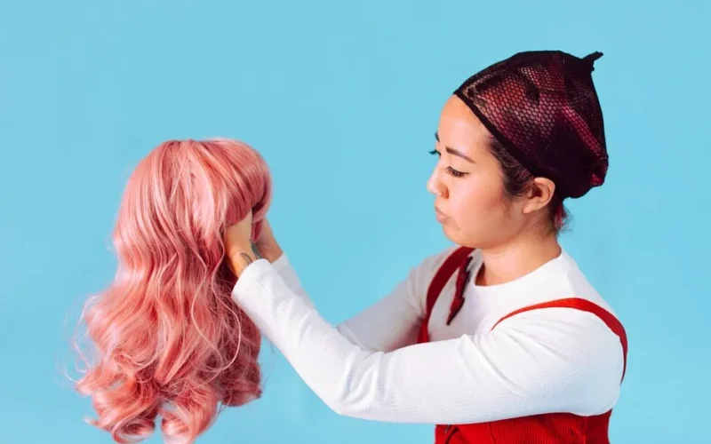 An Asian woman in a net cap holds a pink wig against a blue backdrop, showcasing style and hair fashion.