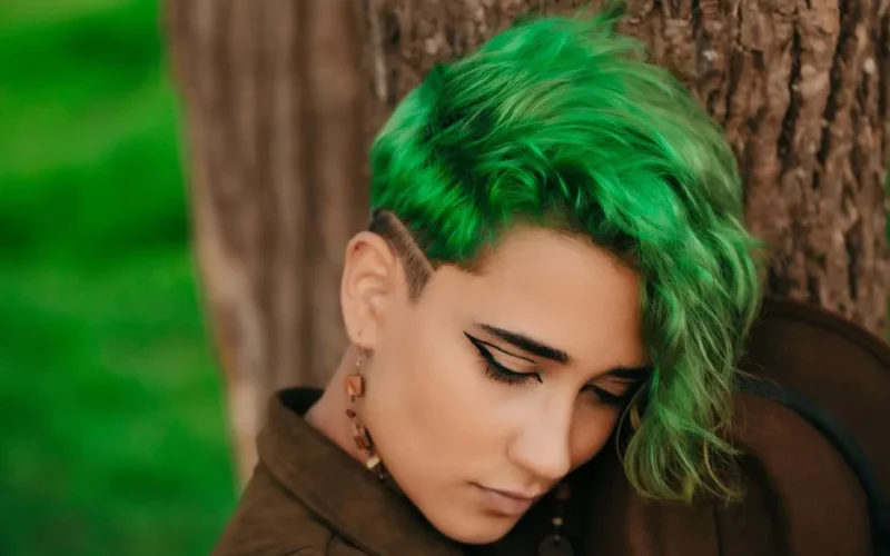 Close-up portrait of a woman with striking green hair posing outdoors against a tree.