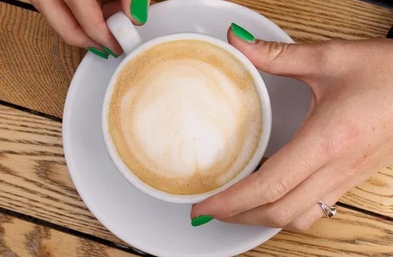 Woman with green nails holds a cappuccino cup on a wooden table.