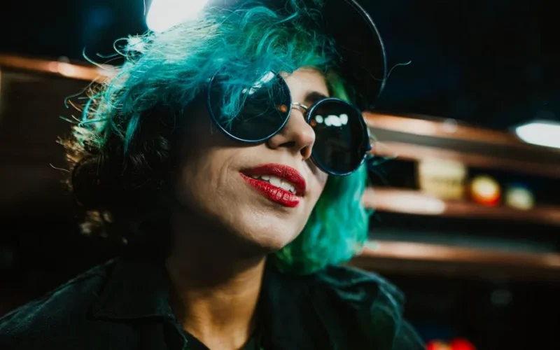 Vibrant portrait of a woman with dyed blue hair, sunglasses, and a cap in a moody setting.