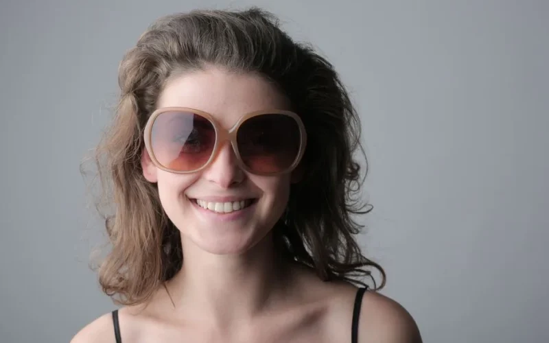 Portrait of a smiling woman with wavy hair and sunglasses against a neutral background.