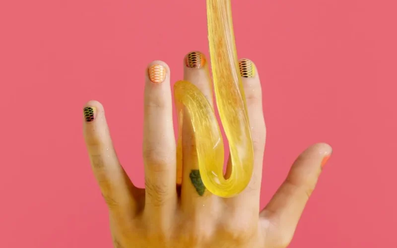 Creative nail art and yellow slime draped on fingers against a pink backdrop.