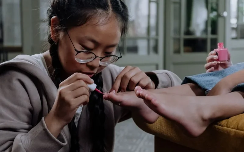Young girl applying pink nail polish indoors, close-up on pedicure with eyeglasses.