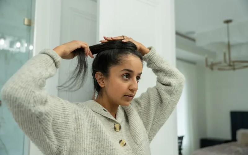 A young woman in a cozy sweater styles her hair into a bun in a bathroom setting.