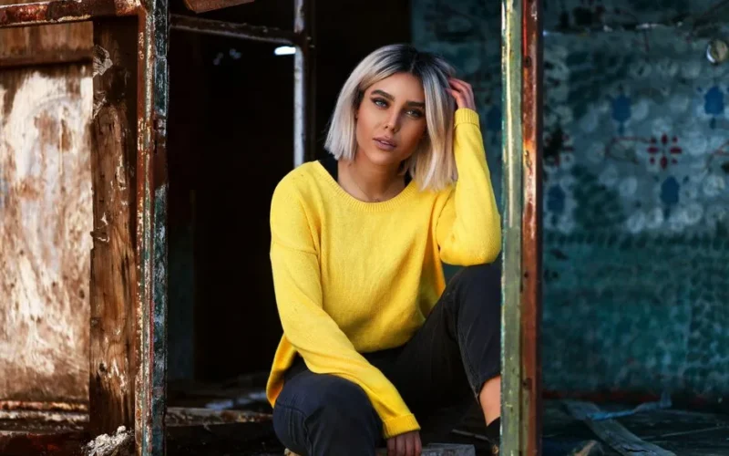 Young stylish female model in colorful clothes touching hair while squatting near shabby wall and looking at camera