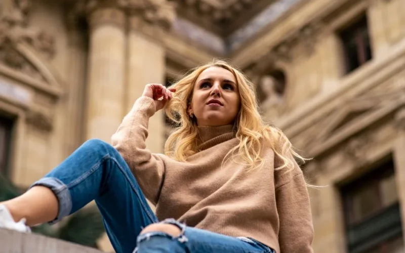 Low angle shot of a woman with blonde hair wearing a beige sweater and jeans, sitting by a historic building.