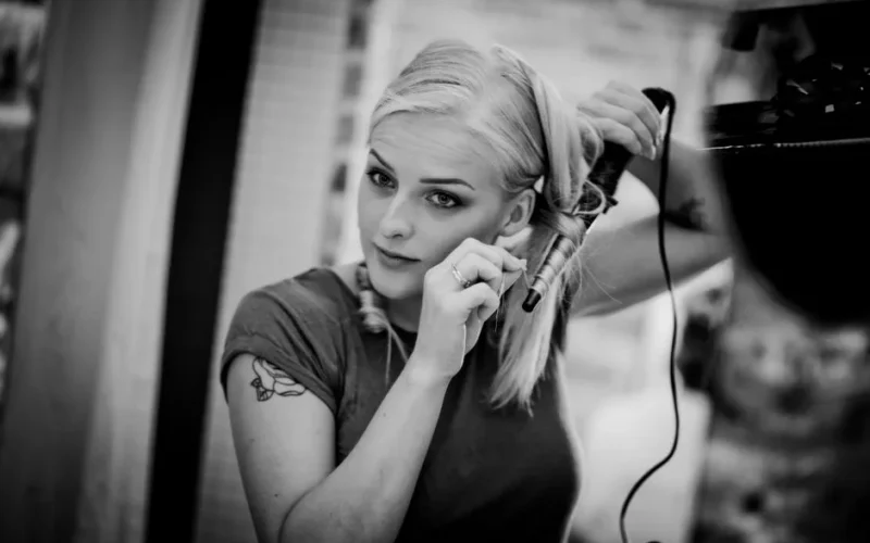 A young woman with tattoos using a curling iron to style her hair in a modern indoor setting.