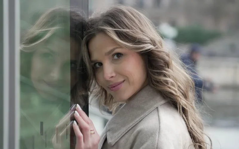 A fashionable woman with brown hair smiling near a window with her reflection in Berlin, Germany.