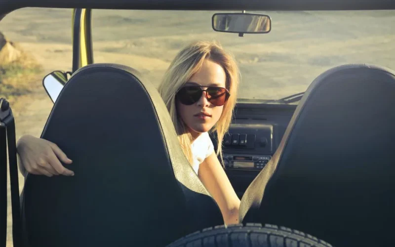 Blonde woman in sunglasses sitting in a convertible on a sunny day, enjoying an outdoor adventure.