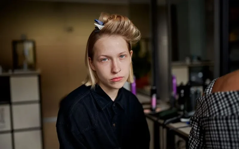 Young unemotional female client looking at camera near crop unrecognizable beauty master in hairdressing salon
