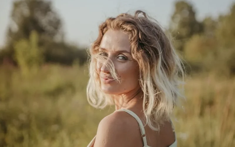 A smiling woman with blond hair glances over her shoulder in a sunlit field.