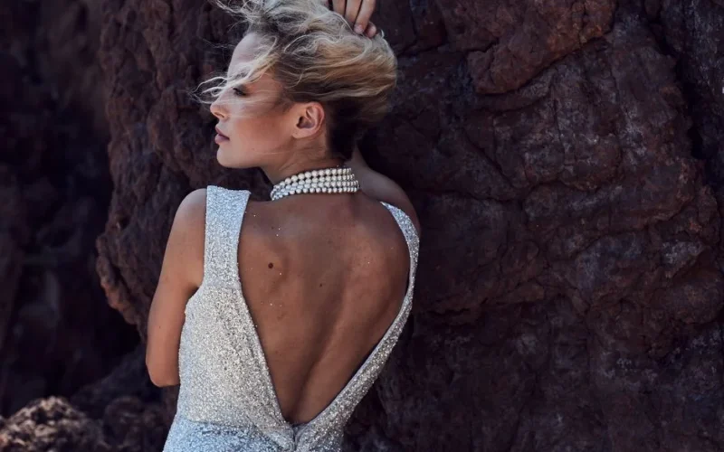 Stylish woman with blonde hair, posing against rocky backdrop in elegant gown and pearl necklace.