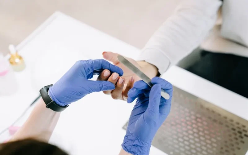 Close-up of a nail technician filing a client's nails with blue gloves in a professional setting.