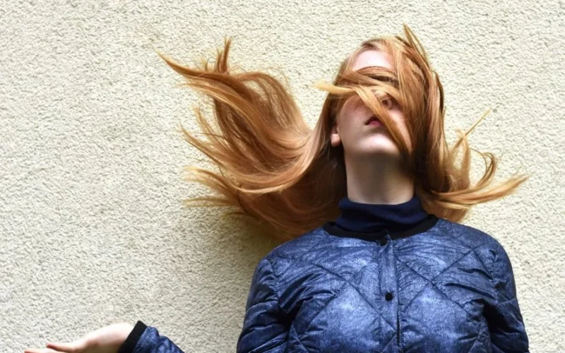 Stylish blonde woman with hair flipping in a bold pose against a textured wall.