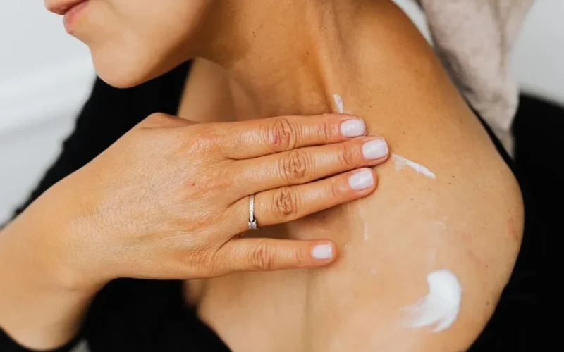 A close-up image of a woman applying skincare lotion on her neck with manicured hands.
