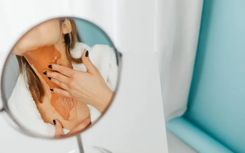 Close-up of a woman applying a thick layer of foundation on her neck in a mirror reflection.