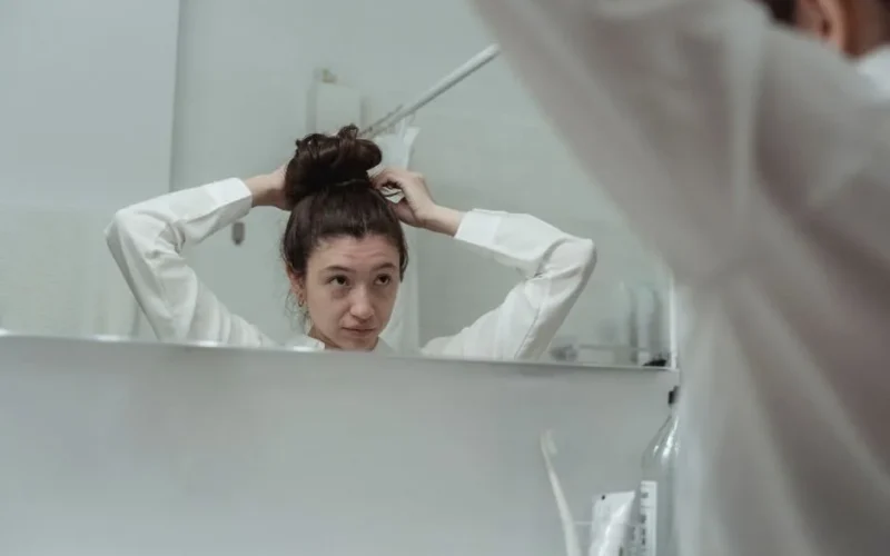 Young woman styling her hair in a bathroom mirror reflection, creating a casual and serene moment.