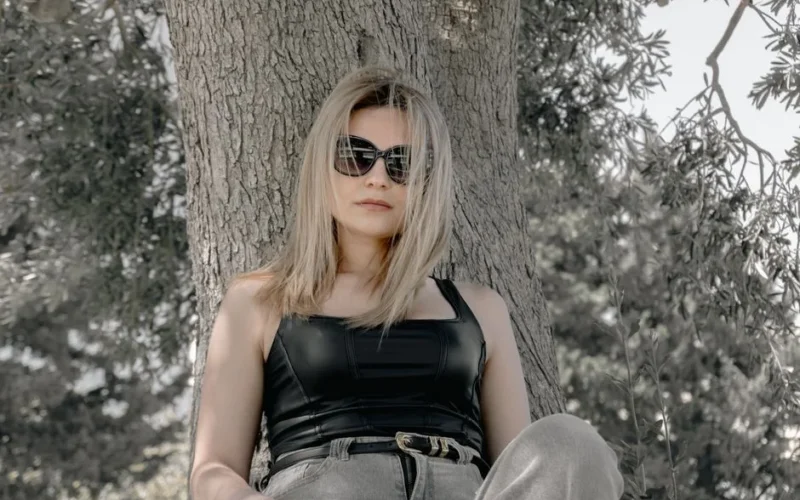 Blonde woman wearing sunglasses and casual outfit relaxing against a tree enjoying a sunny day outdoors.